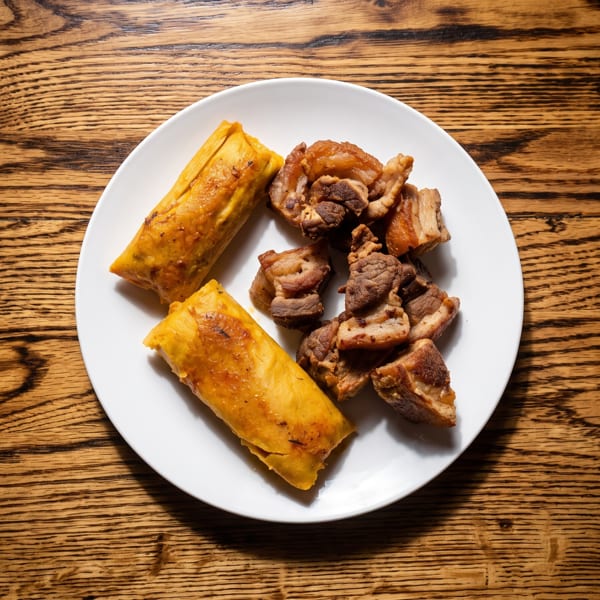 Two golden tamales and a pile of crispy fried pork chunks on a white plate.