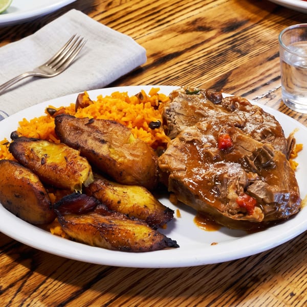 A plate of roasted pork, yellow rice, and fried plantains on a wooden table with a glass of water.