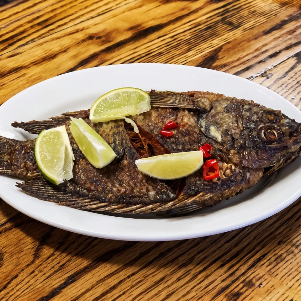 Whole fried fish garnished with lime wedges and red chili peppers on a white plate.